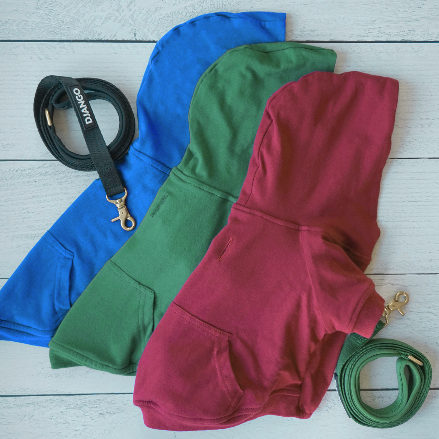 Three different DJANGO dog hoodies laying flat on wood surface showing back pocket details and comfortable fabric